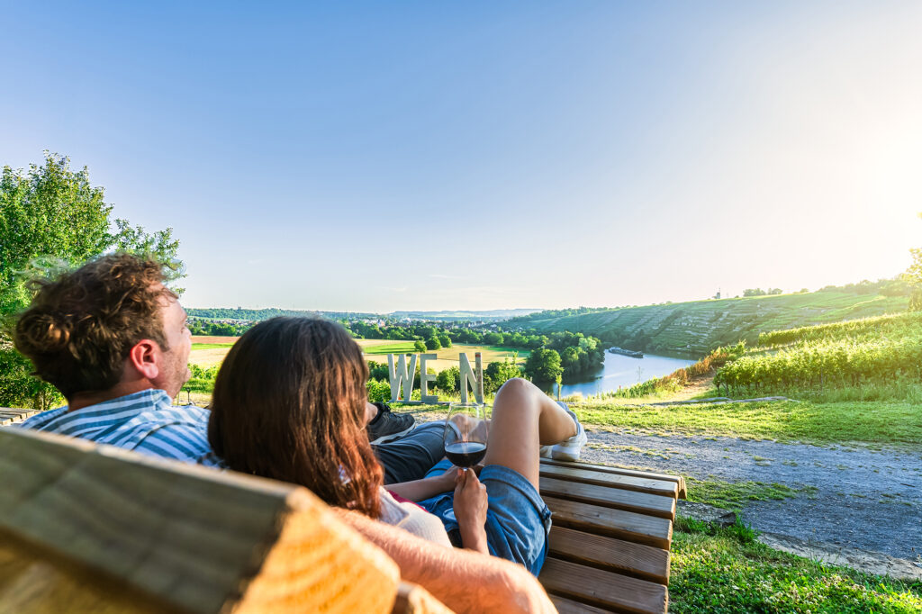 Zwei Personen mit Weingläsern in der Hand schauen in den Sonnenuntergang. Im Hintergrund sind die Weinberge zu sehen.