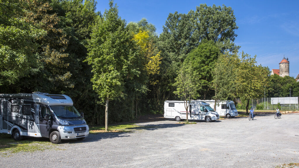 Drei Wohnmobile auf einem Wohnmobilstellplatz. Im Hintergrund sind Bäume und ein Turm zu sehen.