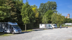 Drei Wohnmobile auf einem Wohnmobilstellplatz. Im Hintergrund sind Bäume und ein Turm zu sehen.