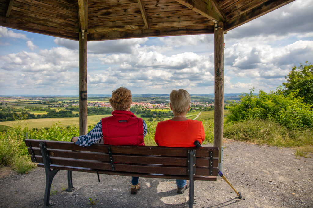 Zwei Personen sitzen auf einer Holzbank unter einem Pavillon und schauen in die Landschaft.