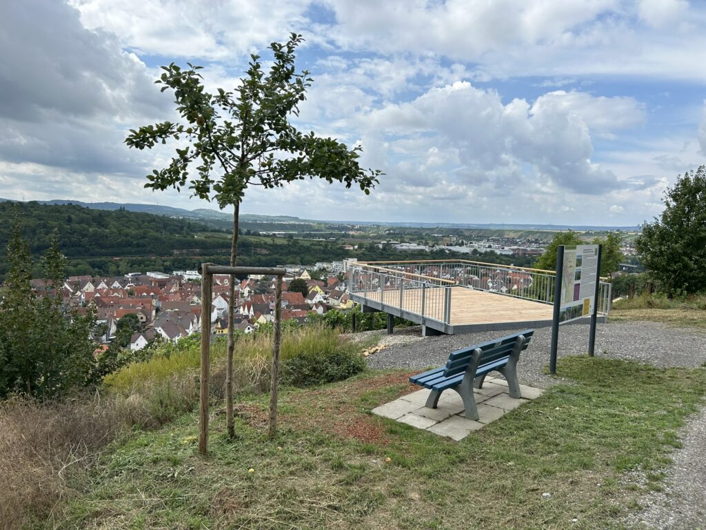 Im Hintergrund sieht man eine hölzerne Plattform, die in die Landschaft ragt. Vorne steht eine blaue Sitzbank.