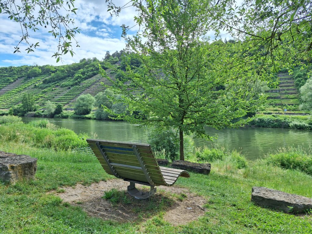 Im Vordergrund steht eine Liege aus Holz, die sich drehen lässt. Im Hintergrund sind ein Fluss und Weinberge zu sehen.